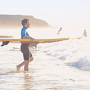 Entrando al agua para tu curso de surf Empieza tu curso de surf en Fuerteventura: con la tabla en la mano te diriges hacia las olas y comienza tu aventura aprendiendo a surfear.